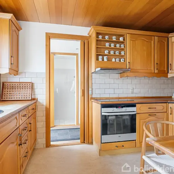 Køkken med træskabe og hvide fliser, inkluderer en ovn og en vaskemaskine. Et bord og stole står i nærheden. Et åbent vindue tilføjer lys til rummet.