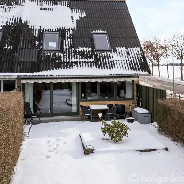 Hus med sne på taget, terrasse foran med havemøbler og planter, omgivet af hække. Vejen ses i baggrunden med marker og træer.