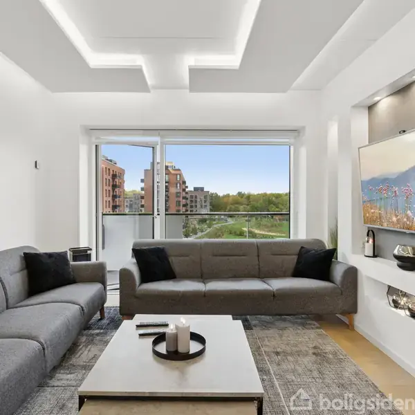 To grå sofaer står i en moderne stue med et sofabord imellem. Store vinduer viser en udsigt over bygninger og træer. Et tv er indbygget i væggen.