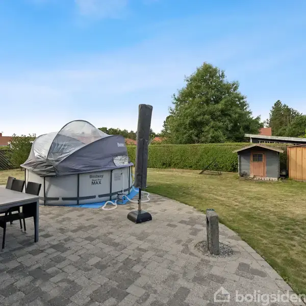 Et oppusteligt badebassin står på en flisebelagt terrasse. Til venstre ses et spisebord med stole, og i baggrunden er en græsplæne med et skur.