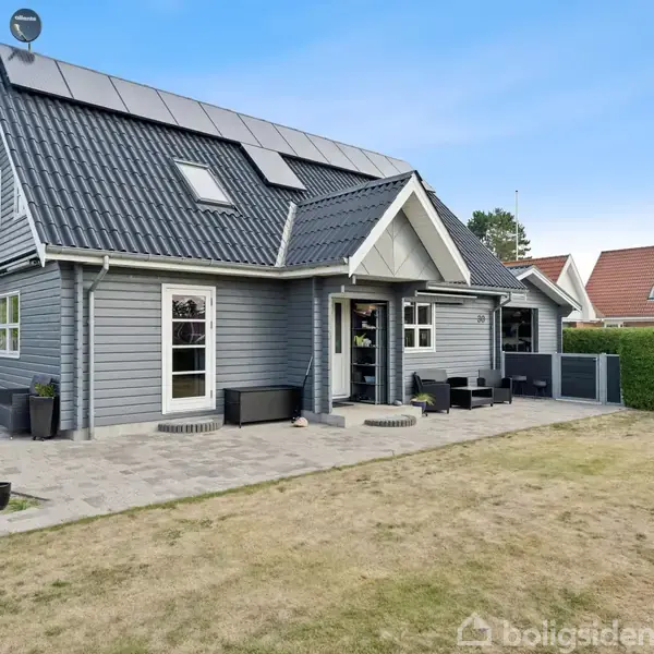 Gråt træhus med solpaneler på taget, omgivet af terrasse og have, står i et beboelsesområde med liggende hus ved siden af. Haveborde og stole er placeret på terrassen.
