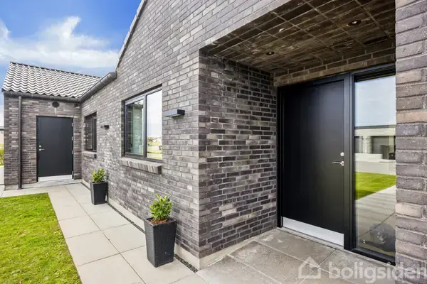 En moderne murstensbygning med sorte døre og vinduer står i en velholdt gårdhave med grøn græsplæne og flisebelægning. To sorte planter med grønne buske flankerer indgangen.