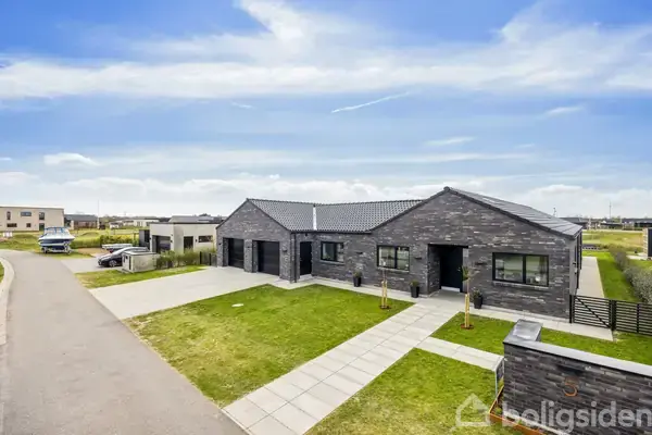 En moderne, grå murstensvilla med sort tag står på en velplejet græsplæne. En flisebelagt gangsti fører til hoveddøren. Villaen ligger i et boligkvarter.