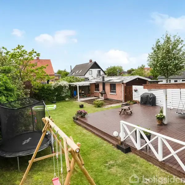 Træterrasse med havemøbler og plantekrukke står ved et rødt murstenshus. En have med en trampolin og gyngestativ omgiver terrassen, og buske samt huse ses i baggrunden.