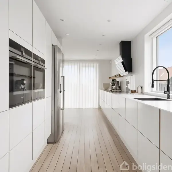 Et moderne køkken har hvide skabe, en indbygget ovn og en stål-køleskab. En stor vask med sort armatur er placeret ved et vindue, som giver naturligt lys i rummet.