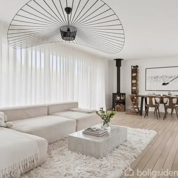 En lys stue med en hvid sofa ved siden af et sofabord. En moderne lampe hænger fra loftet. I baggrunden står et spisebord, og der er en brændeovn.