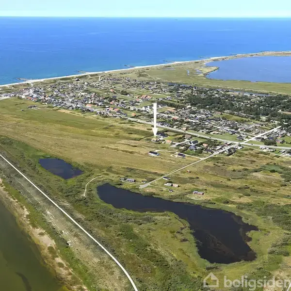 Et landskabsfoto viser en kystlinje med en by i nærheden af vandet. En hvid skorsten står centralt. Området er omgivet af grønne marker, vandløb og flere små søer.