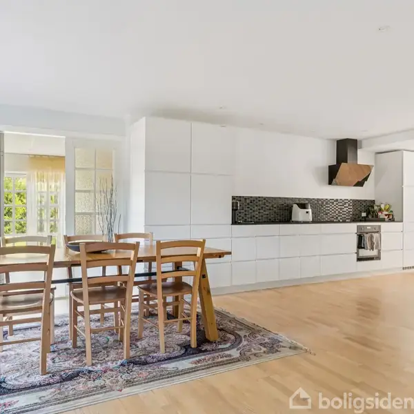 Et lyst køkken-alrum med trægulv, hvor et spisebord med seks stole står på et orientalsk tæppe. Køkkenet har hvide skabe og sort flisebaggardin, mens en sofa er i højre side.