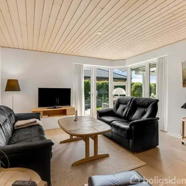 Sort lædersofa og lænestol står i en lys stue med trægulv og træloft. Et sofabord er placeret foran sofaen, og et fladskærms-tv er placeret i et hjørne ved et vindue.