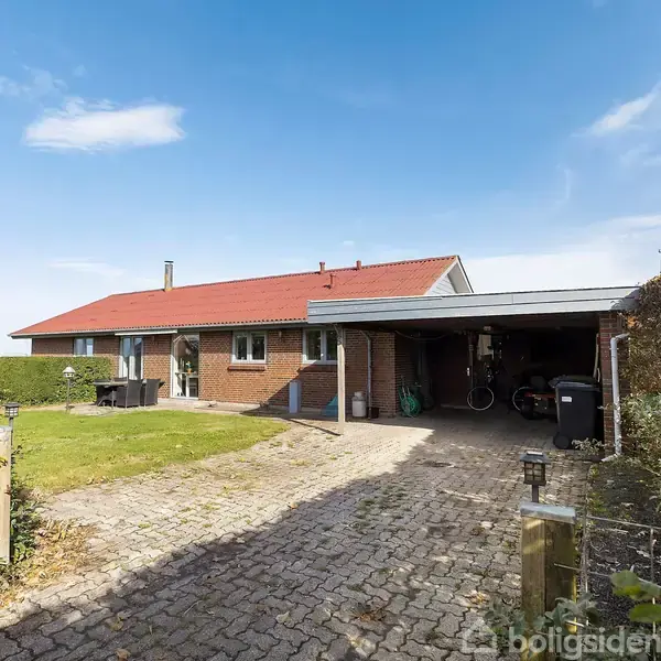 En murstensvilla med rødt tag står stille i et åbent forstadsmiljø. En flisebelagt indkørsel fører til en carport ved huset, omgivet af en velplejet have og hæk.