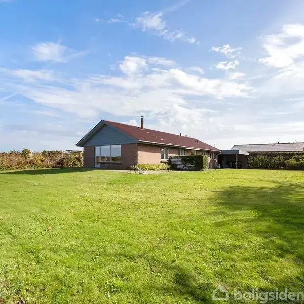 Hus står alene på en stor græsplæne. Skyer bevæger sig på den blå himmel i baggrunden. Omkring huset er der grønne buske og træer.