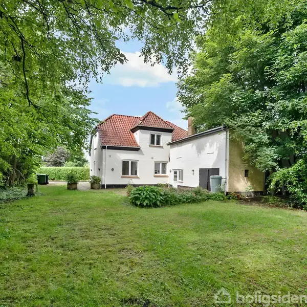 Hvidt hus med rødt tegltag, står stille omgivet af grønne træer og buske i en frodig have. Græsområdet er velplejet og omgivet af tæt skov på alle sider.