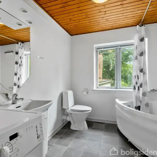 Badeværelse med vaskemaskine, toilet og hjørnebadekar. Træloft og gråt flisegulv. Et vindue med udsigt til natur. Hvide vægge og et spejl over vasken.