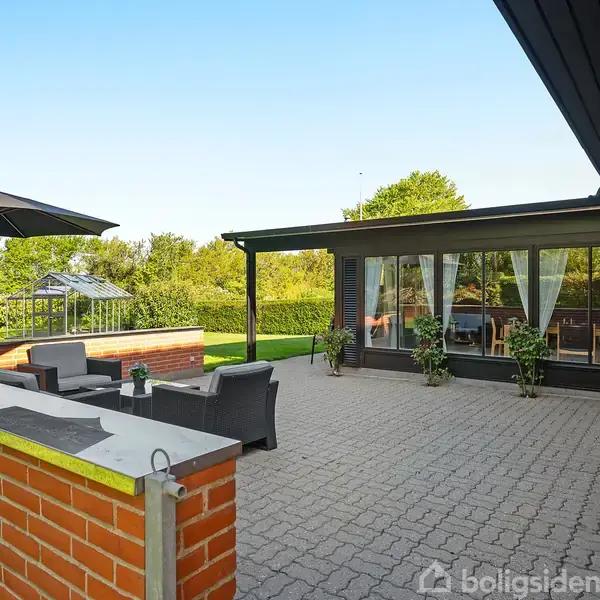 Terrasse med sofaer og parasol står foran et hus med store vinduer og hvid-gardiner. Omgivet af grønt græs og træer, giver opholdsområde udsigt til en fredelig have.