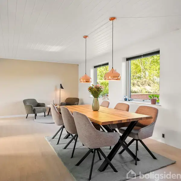 Spisebordet dominerer med seks polstrede stole omkring det. To loftslamper hænger over bordet. Rummet er lyst med store vinduer, der giver udsigt til en grøn have.