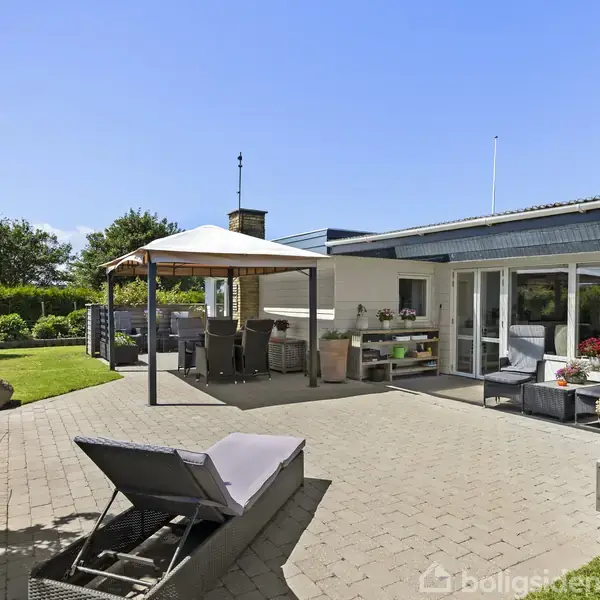 En terrasse med havemøbler og en pavillon står i en solrig have. Omkring er der græsplæne og træer. En liggestol er placeret på fliserne ved huset.