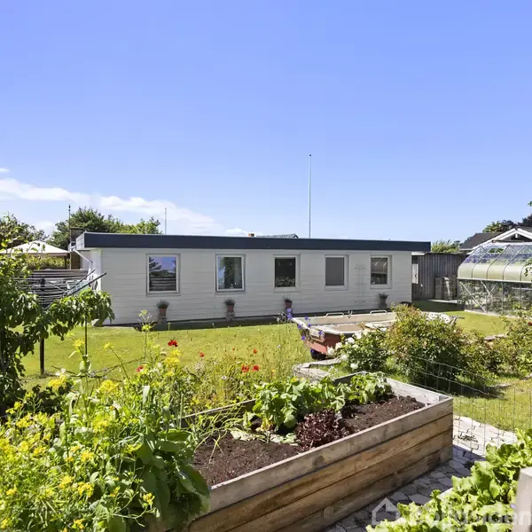 Et hvidt hus står i baggrunden, omgivet af en velplejet have med en stor græsplæne. I forgrunden ses højbede med forskellige planter. Solskin fylder det åbne område.