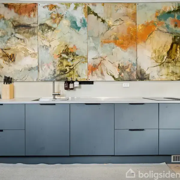 Blå køkkenbord med skuffer under en abstrakt maleri, der dominerer væggen. Maleriet er farverigt med orange, grøn og blå nuancer. Køkkenet indeholder knivblok og sæbedispenser.