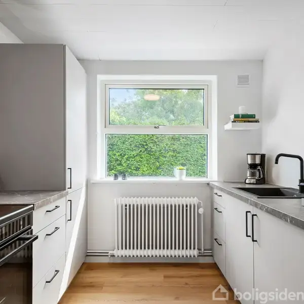 Et moderne køkken med hvide skabe og sort bordplade. En kaffemaskine står ved vasken. Et vindue med udsigt til grønne buske giver naturligt lys. Trægulv og lys vægfarve.