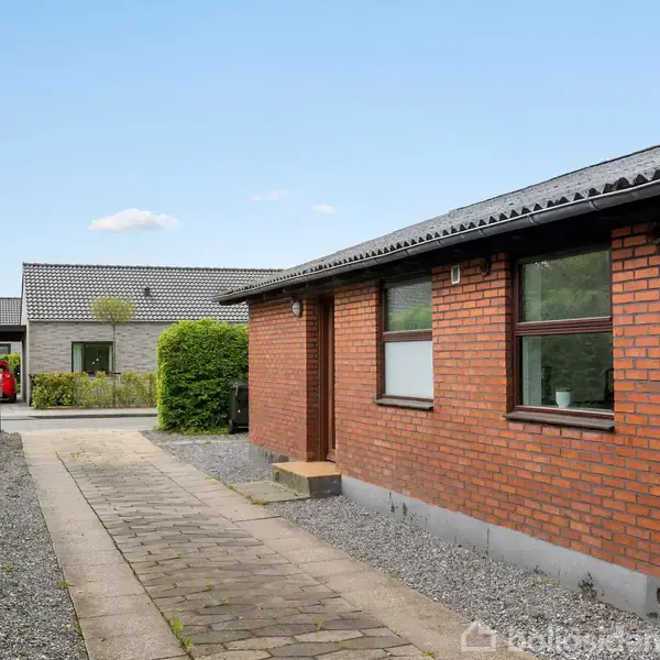 Rødt murstenshus med fladt tag står ved en flisebelagt gangsti. Til venstre er der en tætklippet hæk, og i baggrunden ses en carport og et træhus.
