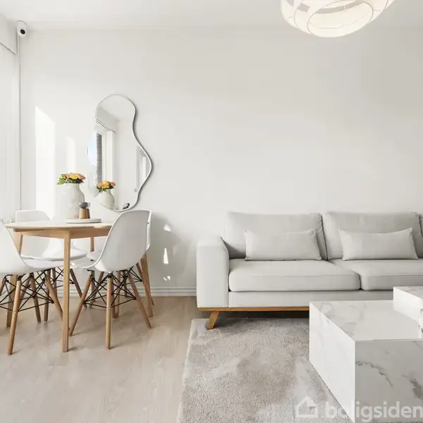 En lys stue med en grå sofa står til højre. Et rundt træbord med stole er placeret ved vinduet. En abstrakt spejl hænger på væggen, og der er blomster på bordet.