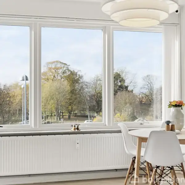 Et bord med fire stole står ved et stort vindue. Sollys strømmer ind og belyser rummet. Udenfor ses træer og en blå himmel. En vase med blomster står på bordet.