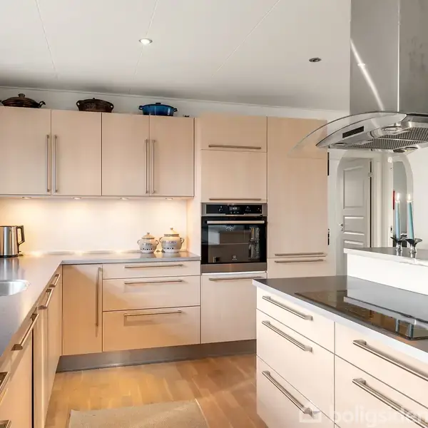 Et moderne køkken med lyse træskabe og integrerede apparater. En kogeø med emhætte centralt. Vindue ved siden af vask giver naturligt lysindfald. Flere dekorative elementer ses på hylderne.