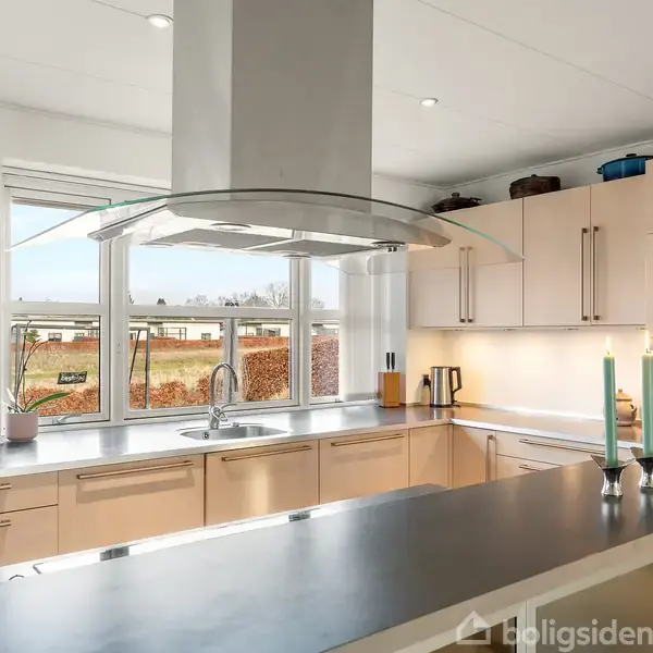 Et moderne køkken med en stor emhætte centralt placeret over en køkkenø. Køkkenet har lyse skabe, dobbelt ovn, og vinduer med udsigt til en have. Kaffemaskine og salt-/pebersæt på bordet.