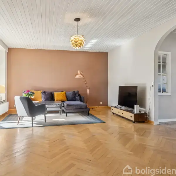 Stue med sofa og stole omkring sofabord på tæppe, tv på lavt bord, træmønstret gulv, brune og grå vægge, bøger på reol og en buet døråbning.