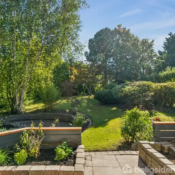 En velplejet have, omgivet af grønne buske og træer, strækker sig ud fra en stenterrasse med trapper. Sollys filtreres gennem trækronerne, skaber en fredelig atmosfære.