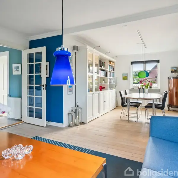 Blåt pendellampe hænger over sofabord med glasdekorationer i lys stue. Blå sofa, hvid reol og spisebord med stole i baggrunden. Vinduer og dør til venstre.