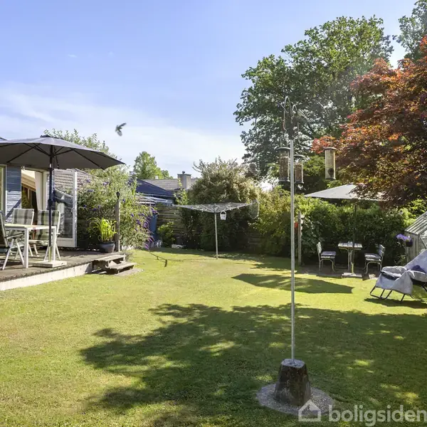Terrassebord med parasol står på verandaen; i baghaven er der en tørresnor, græsplæne, farverigt træ og drivhus omgivet af buske, under en klar blå himmel.