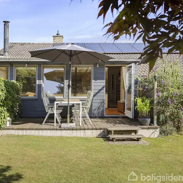 Et lille hus med terrasse, hvor et havebord og parasol står. Baghaven er omgivet af grønne planter og græsplæne, og der ses solpaneler på taget.