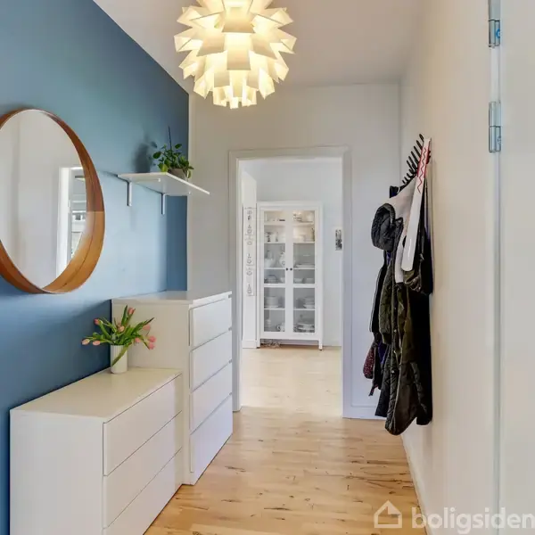 En lys gang med en rund spejl på en blå væg over en hvid kommode. En moderne lampe hænger fra loftet, og der er tøj hængende på højre væg.
