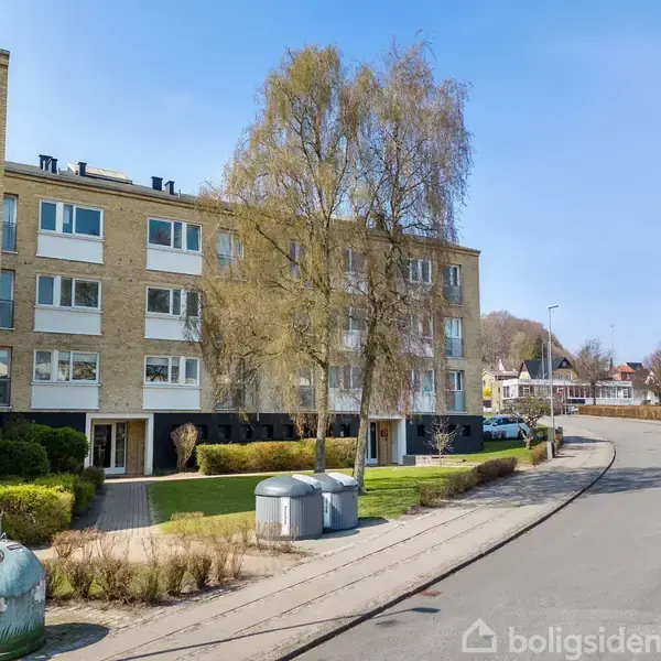 Et treetagers lejlighedskompleks i gule mursten står langs en rolig vej. Foran bygningen ses en grøn affaldscontainer og buske, mens biler er parkeret langs fortovet.