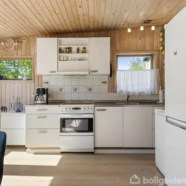 Køkken med trævægge og loft. Komfur og kaffemaskine står på hvidt køkkenskab. Vinduer tillader indstrømmende lys, afslører grøn udsigt udenfor. Køleskab står på højre side.