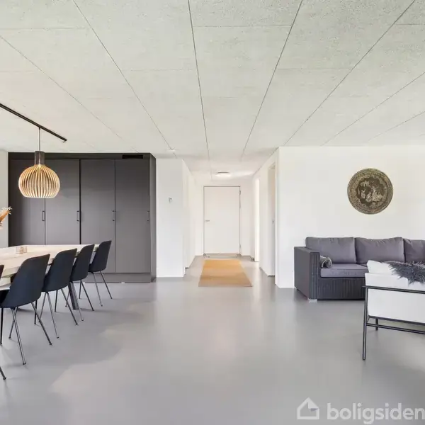 Spisestue sofaen, en lang bord med sorte stole og trælamper i loftet. Til højre en grå sofa og et rundt vægdekoration. Store vinduer giver udsigt til haven.