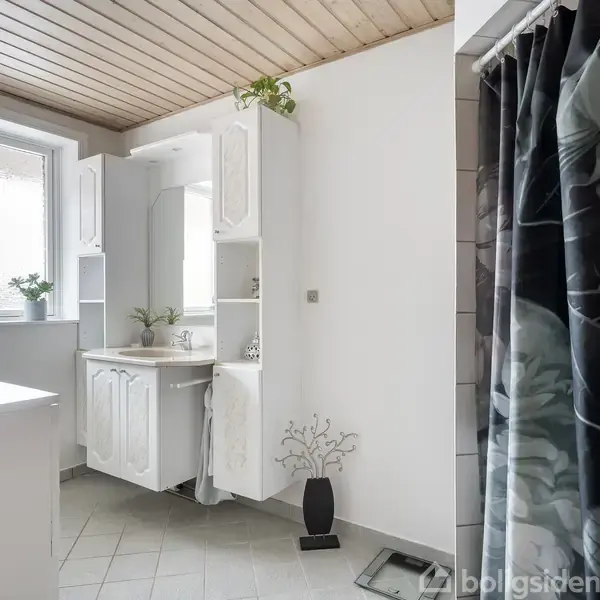 Badeværelse med hvidt skab og vask midt i rummet, grønne planter på skab og vindueskarm. Bruseforhæng til højre af trædør. Flisegulv og træloft.