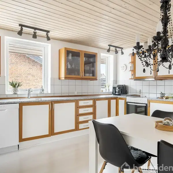 Køkken med hvidt interiør og trækanter, sort lysekrone hængende over hvidt spisebord med sorte stole, omgivet af lyse vægge og store vinduer.