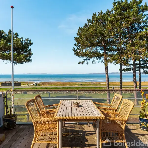Et bord med kurvestole står stille på en terrasse med glasrækværk. I baggrunden ses træer og en udsigt over vandet under en klar blå himmel.