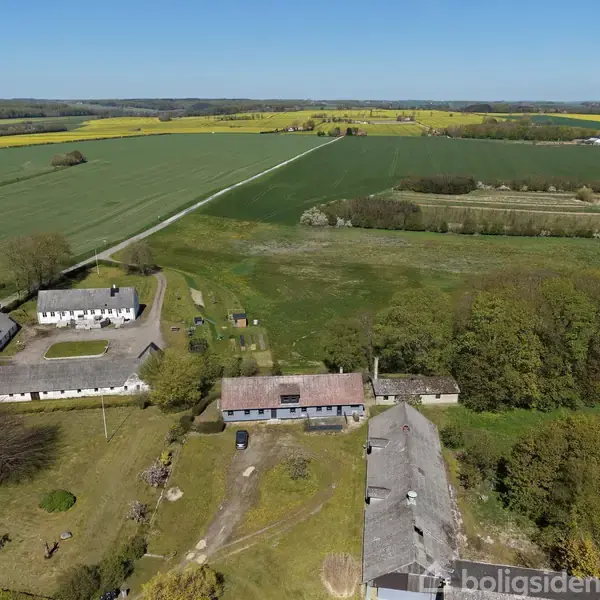 Gårde set ovenfra omgivet af marker og grønne områder; grusveje ses mellem bygningerne. Landskabet strækker sig mod en bred horisont med skove og marker i baggrunden. Ingen synlig tekst.