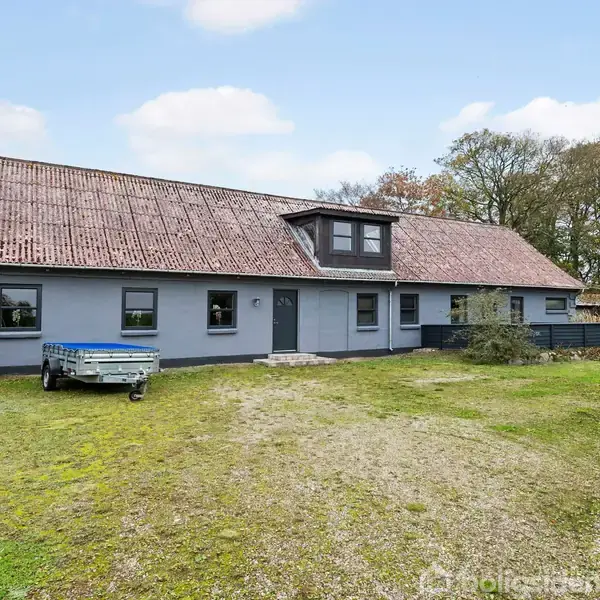 Et stort hus med gråt tag og blågrå facade står stille i en forstadsområde. Græsplænen foran har en lille trailer parket. Træer omkranser stedet i baggrunden.