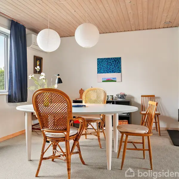 Spisebord med fire stole står i et lyst rum med træloft, ved siden af et vindue med blå gardiner. Pejs i hjørnet, kunst på væggene og planter på vindueskarmen.