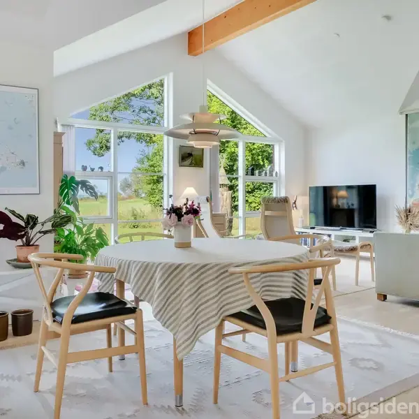 Et lyst, moderne stueområde med et spisebord dækket af en stribet dug og omgivet af to stole. Store vinduer slipper naturligt lys ind, og der er udsigt til grønne omgivelser.