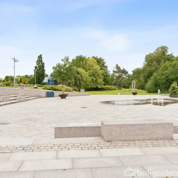 En stor, åben plads med granitbelægning har flere fontæner, der sprøjter vand opad. Omgivet af træer og grønne områder, skaber pladsen et offentligt byrum.