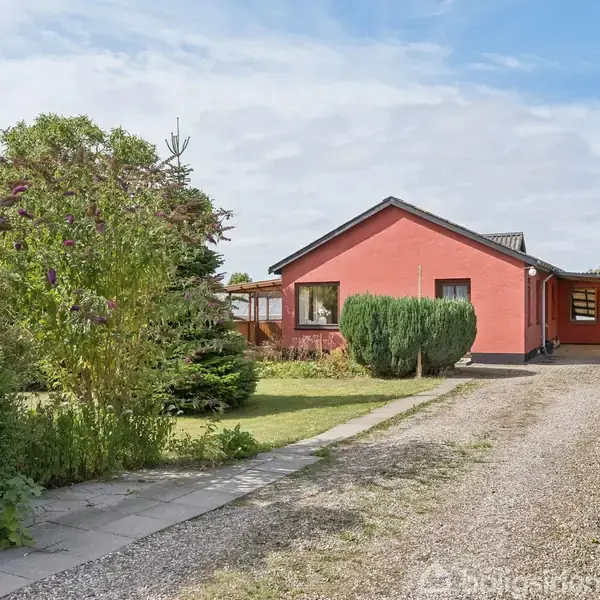 Et rødt hus står stille i en frodig have med grusindkørsel og grønne buske rundt omkring. Himmel og spredte skyer ses i baggrunden.