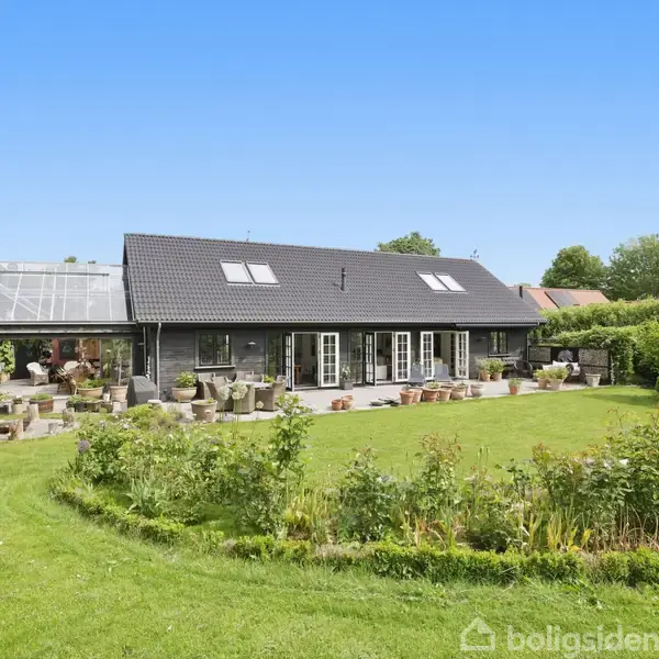 Et moderne hus med sort tag står i forgrunden med flere tagvinduer. En stor terrasse med potteplanter omgiver huset. Baggrunden er en velplejet græsplæne og buske.