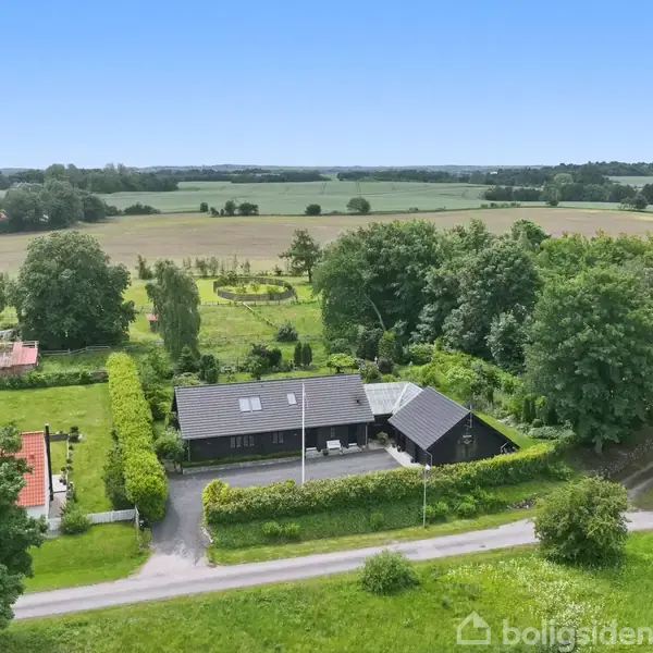 Et L-formet hus med sort tag og glasdrivhus udvidelse befinder sig i en frodig have med buske og blomster. Omkringliggende landskab er grønt og skovklædt.