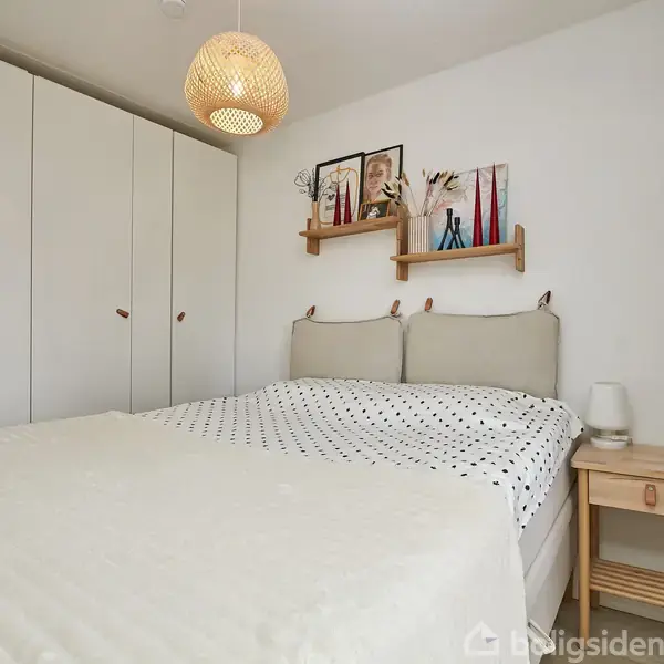 Seng er reddet med prikket sengetøj, står i soveværelse med hvid garderobe og natbord. Væglamper og dekorationer findes på en hylde over sengen.