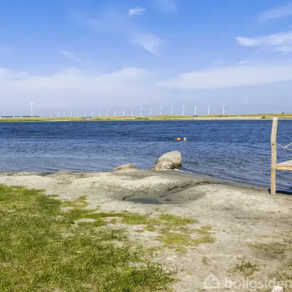 En træbrygge strækker sig over en strand ved en rolig sø. I baggrunden ses vindmøller på en landtange under en klar himmel.